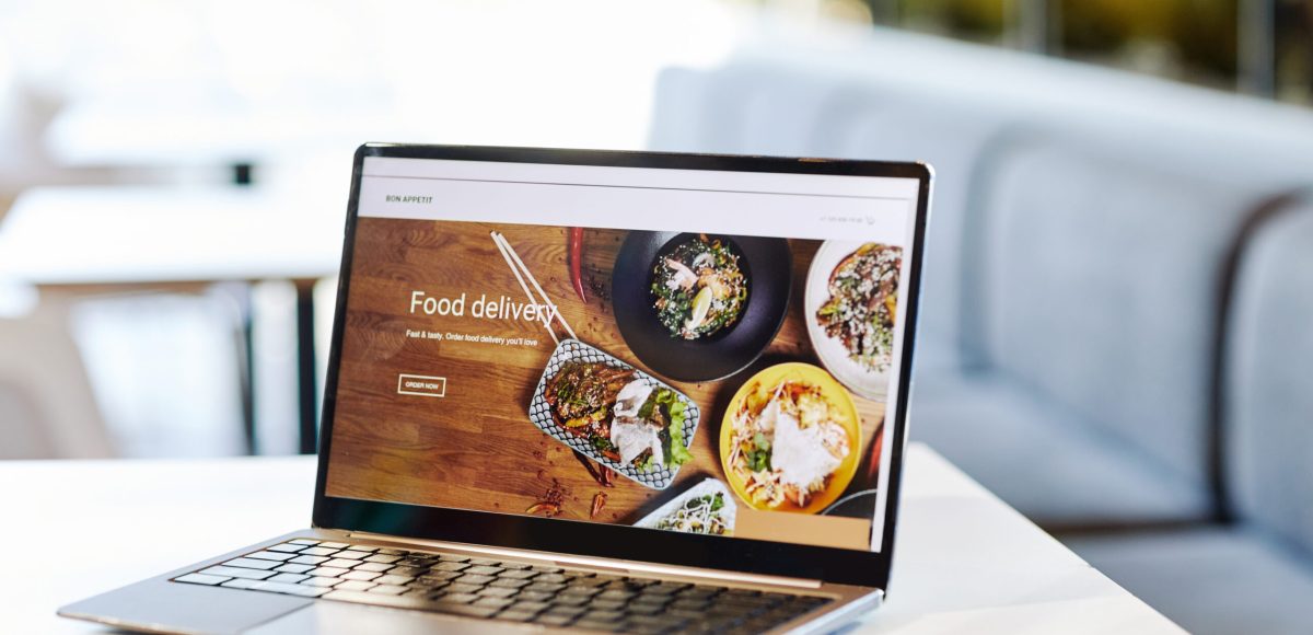 Background image of opened laptop with Food delivery website on table in food court or cafe interior, copy space